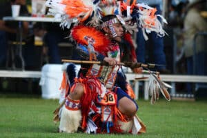 performers wearing vibrant traditional attire perform a dance at a 4th of July powwow, while spectators watch.