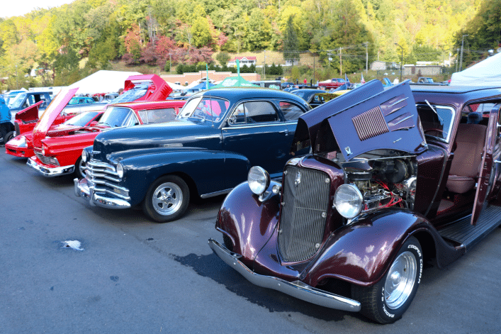 A rod run featuring various vintage cars, including a dark blue and a burgundy vehicle with hoods open, displaying engines in a sunny outdoor setting with trees.