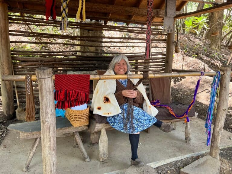 A smiling elderly woman sits on a rustic wooden swing, holding a flatbread, surrounded by colorful woven textiles and a basket, in the quaint outdoor setting of Oconaluftee Indian Village.