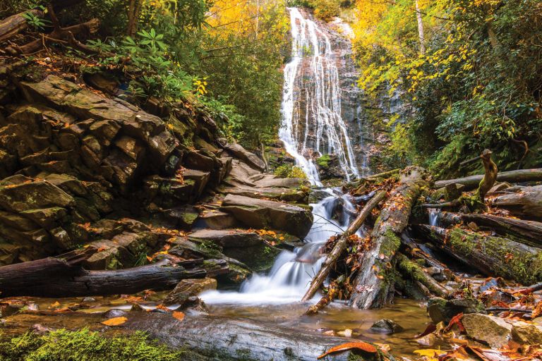 A scenic view of a waterfall cascading down a rocky cliff surrounded by autumn foliage and a small pool at the base in a Cherokee forest.