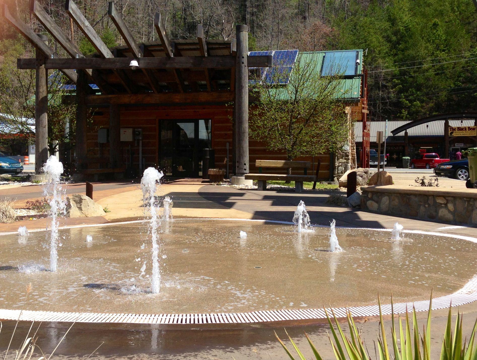 A sunny plaza with a circular Cherokee fountain spraying water, trees, and a rustic building with solar panels on the roof in the background, located in a small town setting.
