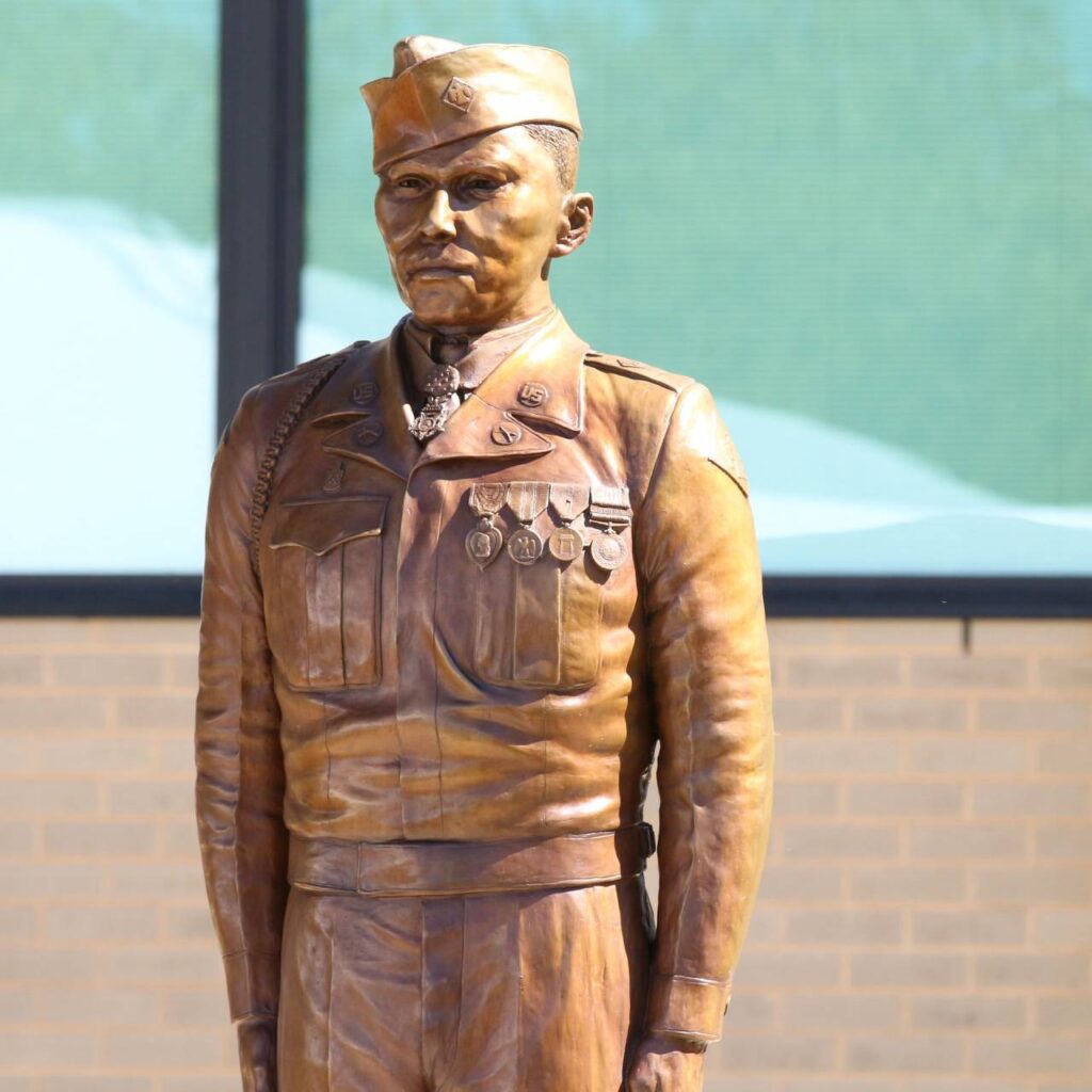 A bronze statue of War Hero Charles George, a military officer in uniform adorned with medals, standing upright against a building with a green window. The officer depicts a stern expression and a formal posture.