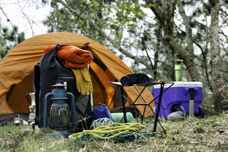 A well-equipped camping site featuring an orange tent, a backpack with a hanging towel, a lantern, a folding chair, binoculars, a rope, and a cooler in a natural, wooded setting