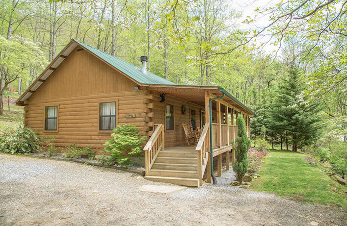 A cozy log cabin with a covered porch nestled among lush green trees in a serene wooded area. A gravel pathway leads up to the wooden steps of the cabin.