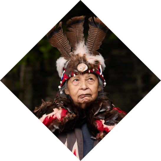 An elderly indigenous person wearing a detailed feathered headdress looks solemnly forward, set against a blurred natural background amid the setting of "Unto These Hills." The headdress includes red and white accents