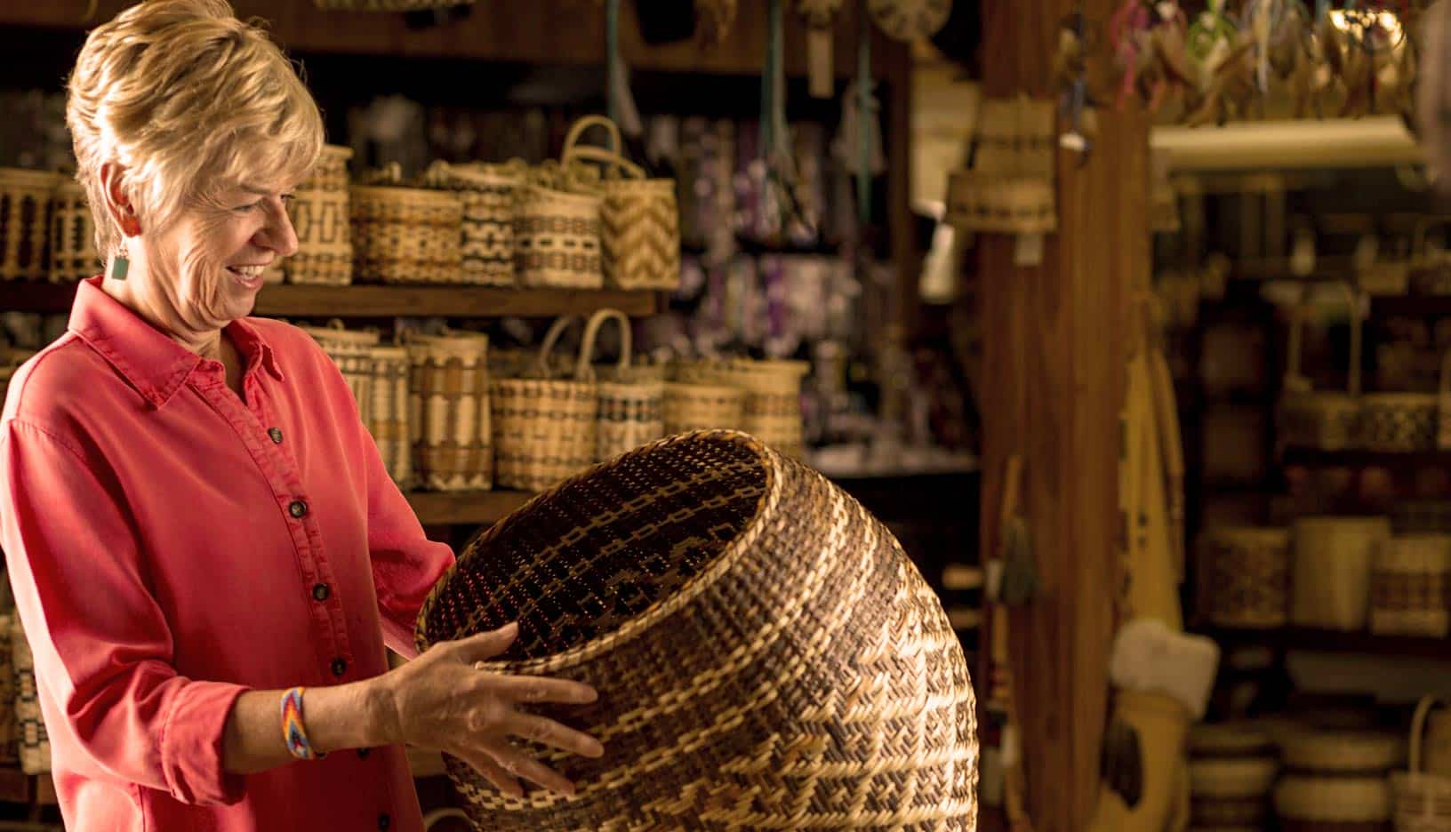 An elderly woman in a red blouse smiles while examining a large woven basket in a store filled with various baskets at the theater-themed market section.