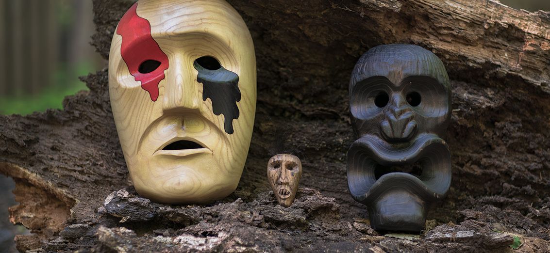 Three unique masks—a painted theatrical mask, a small tribal mask, and a stylized gorilla mask—arranged on a fallen tree trunk in a forest setting.
