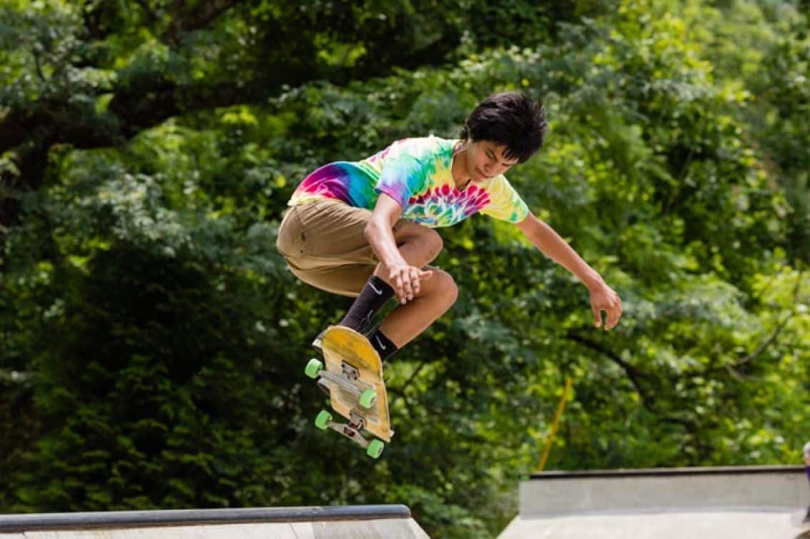 A young skateboarder, wearing a colorful tie-dye shirt, performs a jump trick above a concrete ramp at Cherokee Action Sports Park, surrounded by lush green trees.