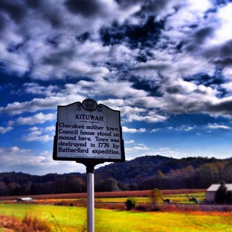 A historical marker titled "Discover Kituwah" with text about its significance as the Cherokee Mother Town. The background features a vibrant sky and a pastoral landscape.