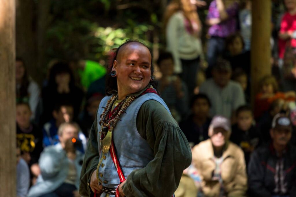A smiling man dressed in traditional native American attire performs outdoors before an attentive audience at Oconaluftee Indian Village in a forest-like setting.