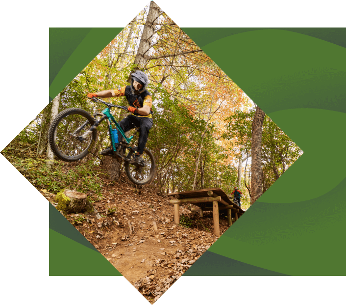 A mountain biker performs a jump on a forest trail, surrounded by autumn-colored trees. the cyclist, wearing a helmet and protective gear, is airborne above a wooden ramp.