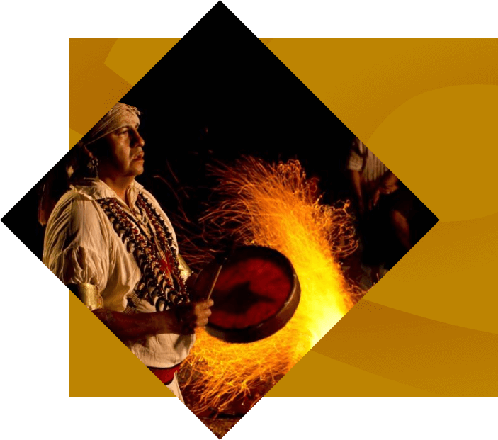 A vibrant bonfire burns brightly in a dark setting, flames twisting and turning against the dim, shadowy background, capturing the essence of Cherokee seasonal attractions while man drums