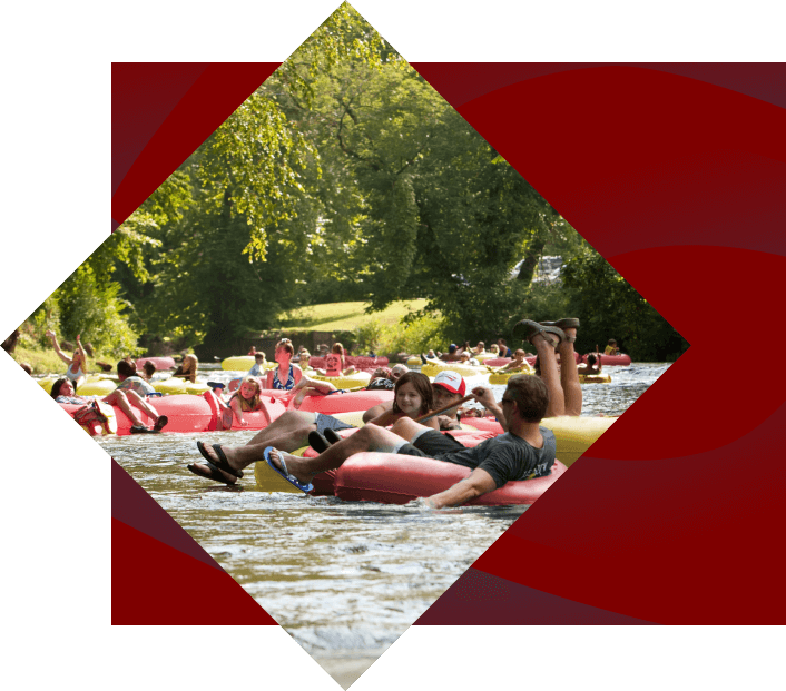 A group of people enjoy a sunny day participating in the outdoor activity of river tubing, floating downstream in colorful inflatable rafts. The setting is vibrant with greenery on the riverbank.