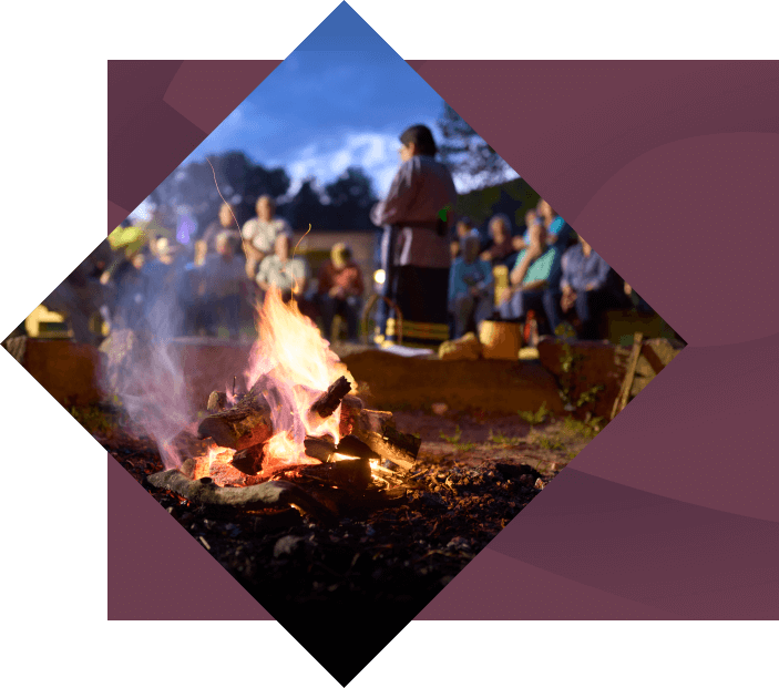 A diamond-shaped image of people gathering around a campfire at dusk, with a woman in sharp focus roasting a marshmallow, and others blurred in the background, conveying a sense of communal culture.
