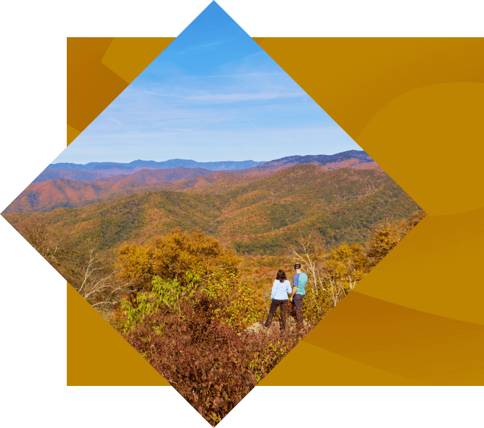 A couple stands together on a mountain, overlooking a vast landscape of colorful autumn trees under a clear blue sky. the image is presented in a diamond shape with a yellow border.