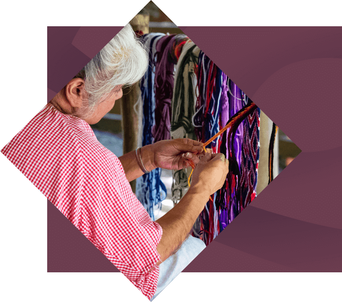 An elderly woman with gray hair, wearing a red and white shirt, sits by a clothes rack in a serene, shaded outdoor setting as she expertly crafts colorful textile art with playful designs.
