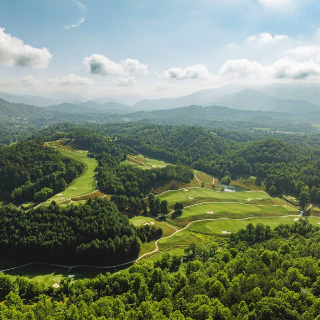 Aerial view of a lush green golf course nestled among rolling hills with a backdrop of hazy mountains and scattered clouds in the sky, showcasing one of the popular Cherokee activities.