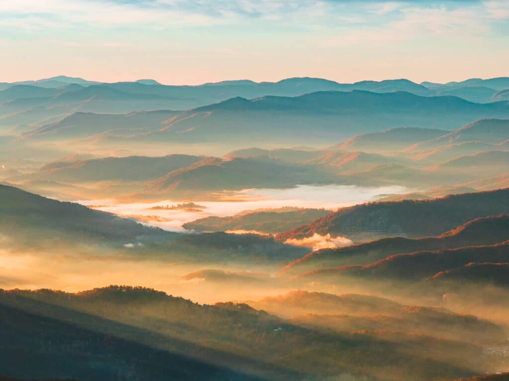 Early morning light illuminates layers of misty mountains and valleys in the Smoky Mountains, with a golden glow covering the landscape and a river meandering through the fog.