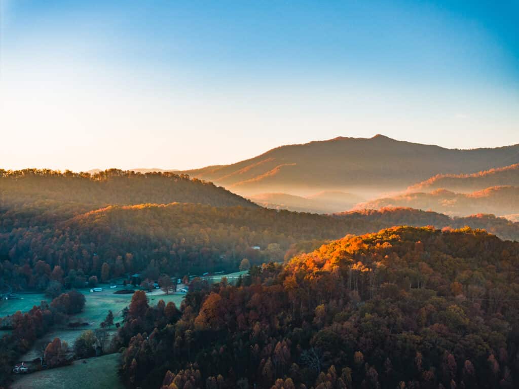 Aerial view of a misty landscape at sunrise featuring rolling hills covered in autumn-colored trees with Sequoyah National Golf Course and a mountain in the background under a clear sky.
