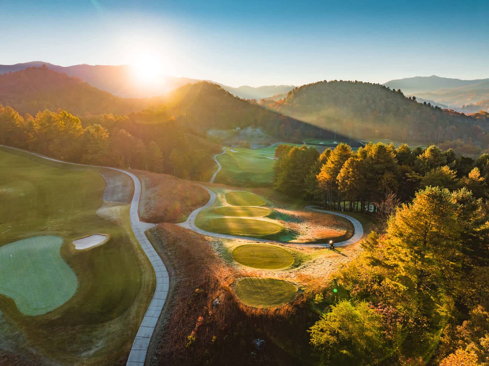 Aerial view of Sequoyah National Golf Course at sunrise, showcasing vibrant autumn trees, undulating greens, and a winding cart path amidst a hilly landscape.