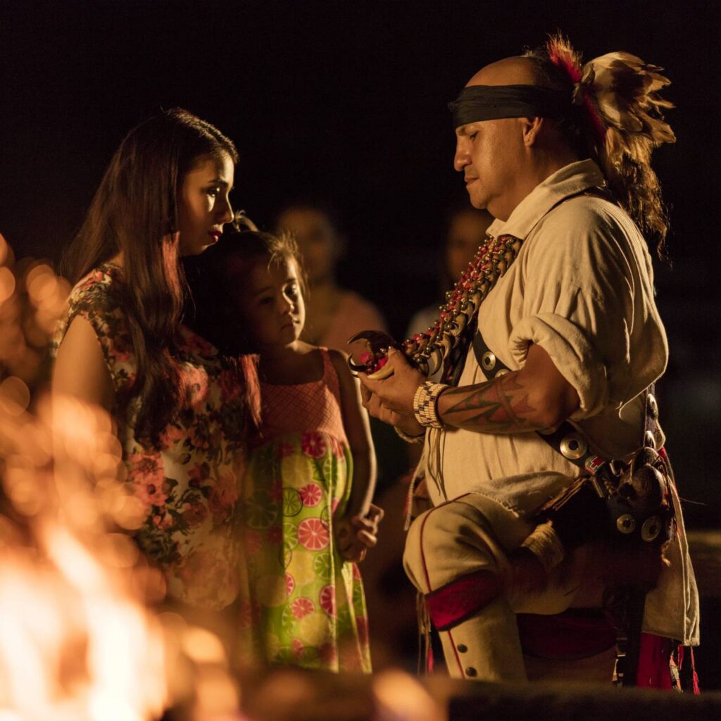 A man in traditional Cherokee attire speaks to a woman and a young girl by a fire at night, conveying cultural heritage.