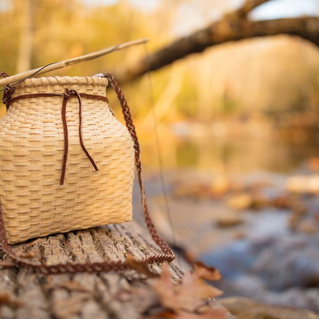 A wicker basket sits on a tree stump by a serene river in Cherokee, surrounded by fallen autumn leaves, with a soft-focus background highlighting warm golden hues of the setting sun.