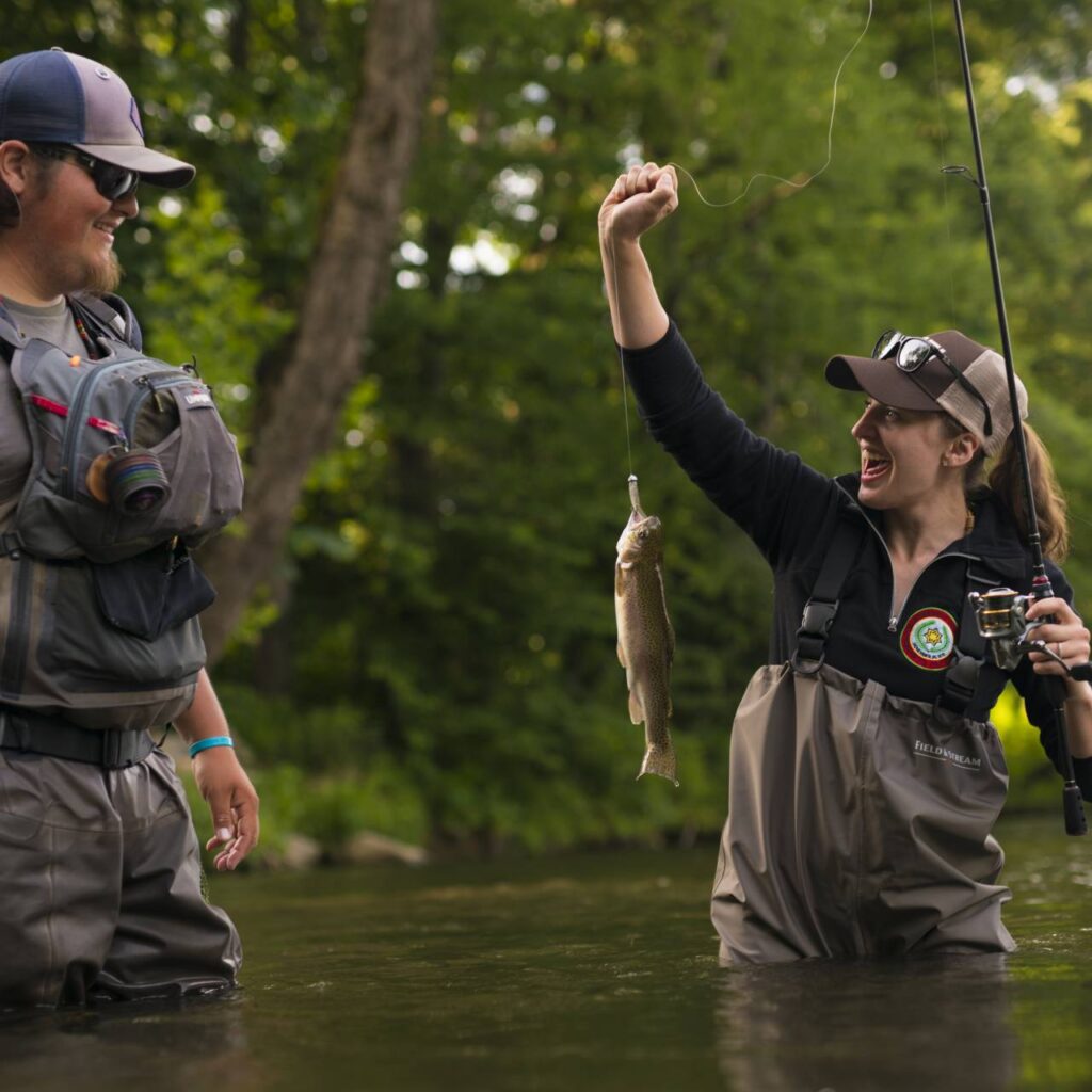 A woman in waders excitedly holds a small fish on her fishing line, while a man in waders looks on with a smile, both standing in a river surrounded by greenery amidst the vibrant