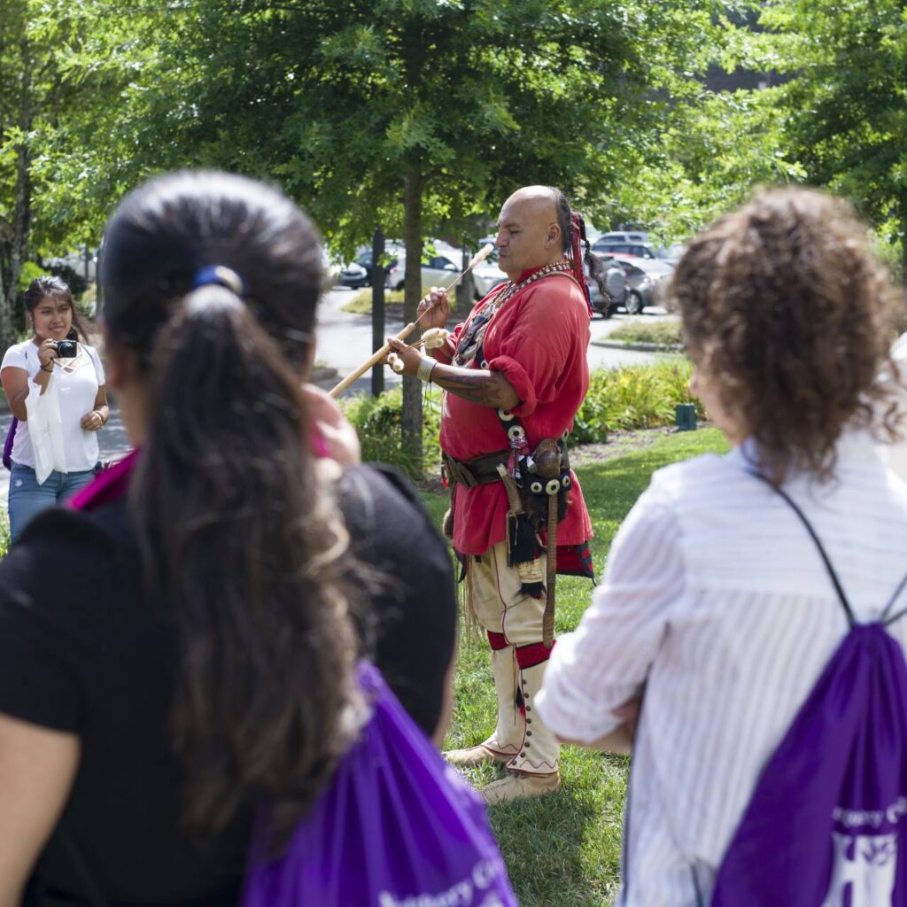 A person in traditional Cherokee attire plays a flute for an attentive audience in a park, surrounded by trees and sunlight, with some listeners holding purple bags.