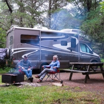 Two people stay by a campfire in front of a parked RV in a forest area, with smoke rising from the fire and lush green trees in the background.
