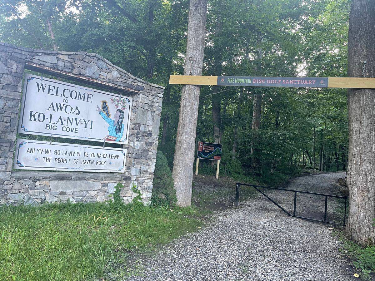 A welcoming sign for the Fire Mountain Disk Golf Sanctuary at the entrance of a wooded trail, with another sign reading "the human disc golf sanctuary" beside it.