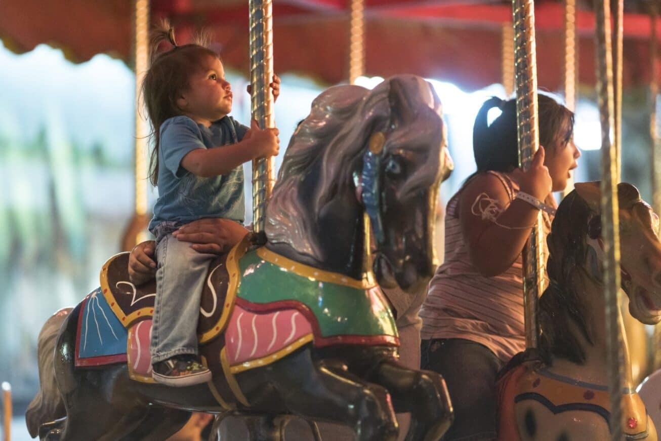 A young child with a joyful expression rides a carousel horse at the Cherokee Indian Fair Grounds, holding onto the pole, as an adult sits beside on another horse, both illuminated by warm, ambient light