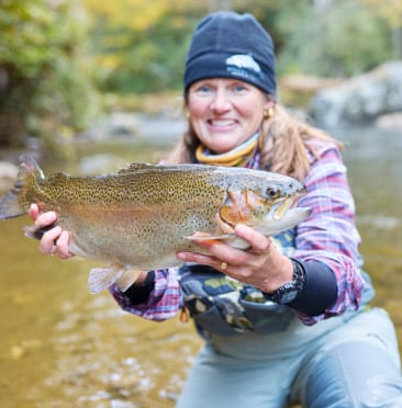 A woman in a beanie and outdoor wear smiling proudly as she holds a large trout in both hands, standing in a river surrounded by autumn foliage.