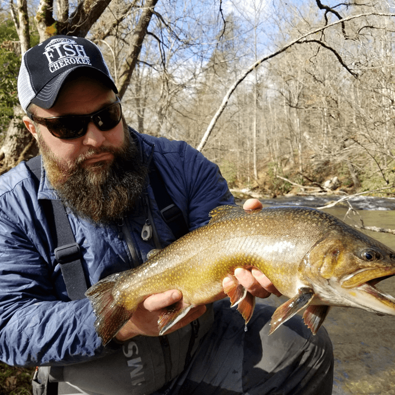 A man with a beard wearing a cap and sunglasses holds a large fish beside a river, showcasing his proud catch in natural daylight.