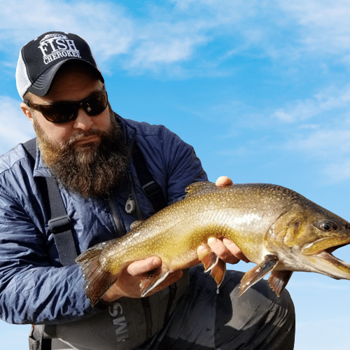 A man with a beard wearing a cap and sunglasses holds a large fish beside a river, showcasing his proud catch in natural daylight.