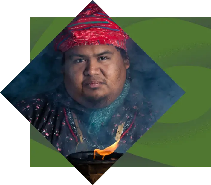 Portrait of a man wearing traditional attire from "Unto These Hills" including a red patterned hat, framed by a diamond-shaped border with green background. He appears serene and dignified.