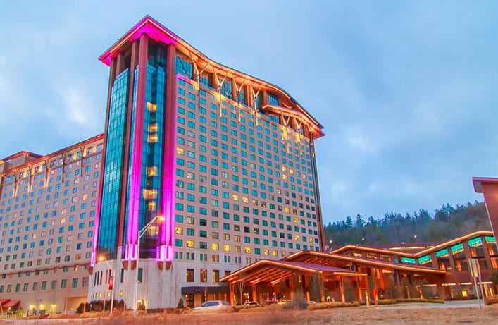 A large modern hotel, Harrahs Casino Resort, with a multi-story facade illuminated by vibrant pink and blue lights, set against a twilight sky, surrounded by hilly terrain.