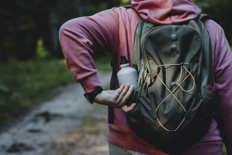 A person wearing a pink hoodie and a backpack holds a water bottle, standing on a forest trail.