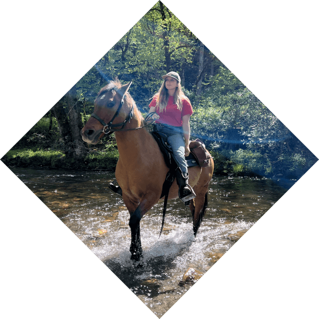 A woman in a red shirt and a cowboy hat riding a brown horse through a shallow, sunlit stream surrounded by green forest during Cherokee spring, presented in a diamond-shaped frame.