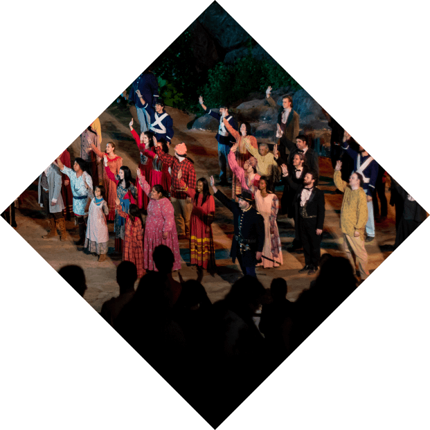 A theater cast receiving applause on stage, viewed through a diamond-shaped frame, during a performance of "Unto These Hills." The scene shows actors in colorful costumes against a naturalistic set with rocks