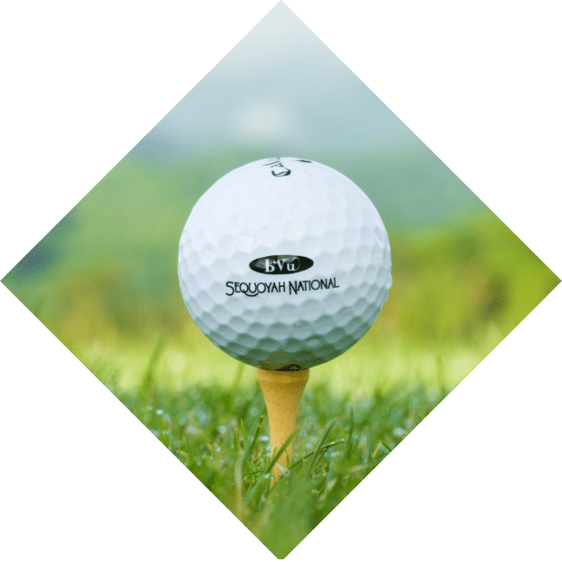 A close-up of a white golf ball labeled "Cherokee Spring" on a yellow tee, set against a soft-focus background of green grass and distant hills.