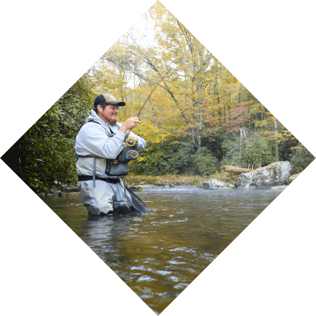 A man in waders stands fishing in a river, surrounded by forested banks in autumn. he is focused on casting his line, holding a fishing rod and reel.