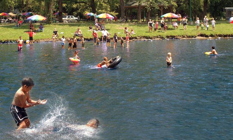 People enjoying a sunny day at Cherokee Island Park with a swimming area. Some are wading and swimming in the water, while others relax on the grass and under umbrellas.