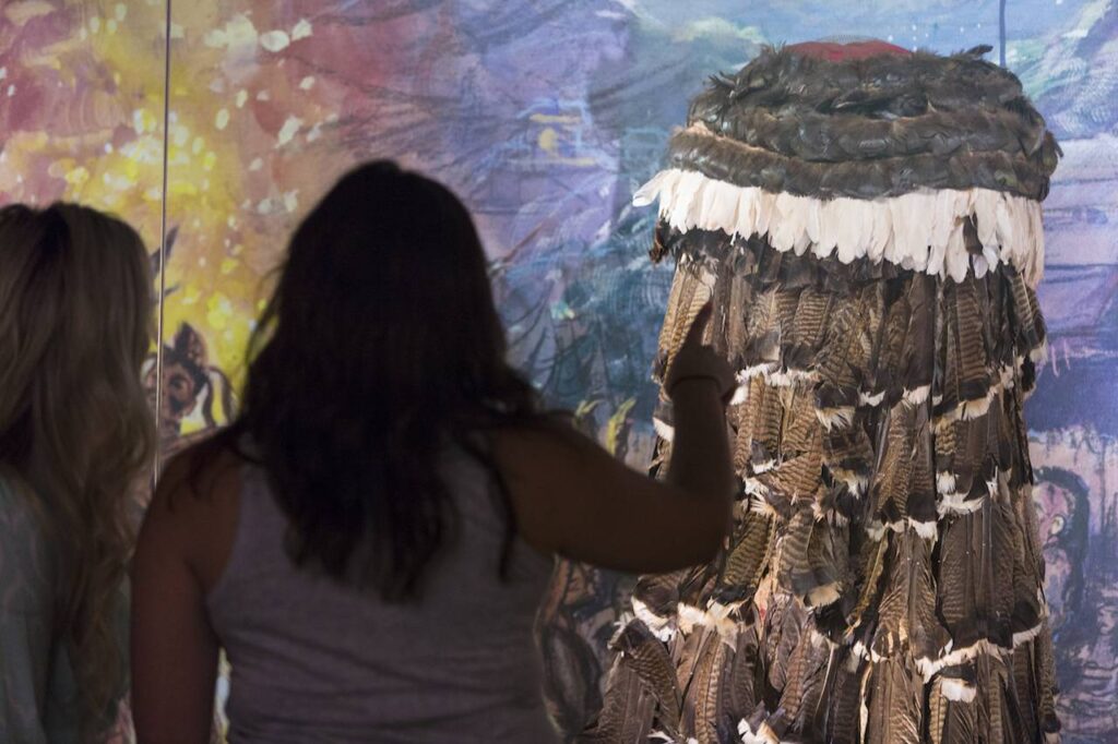 Two individuals viewed from behind, observing a large artistic installation featuring feathers at a Cherokee fall activities exhibition. The background shows a colorful abstract painting.