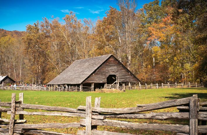 A rustic wooden barn with a large open doorway stands amid lush autumn foliage at the Mountain Farm Museum. A traditional split-rail fence encloses the area under a clear blue sky.