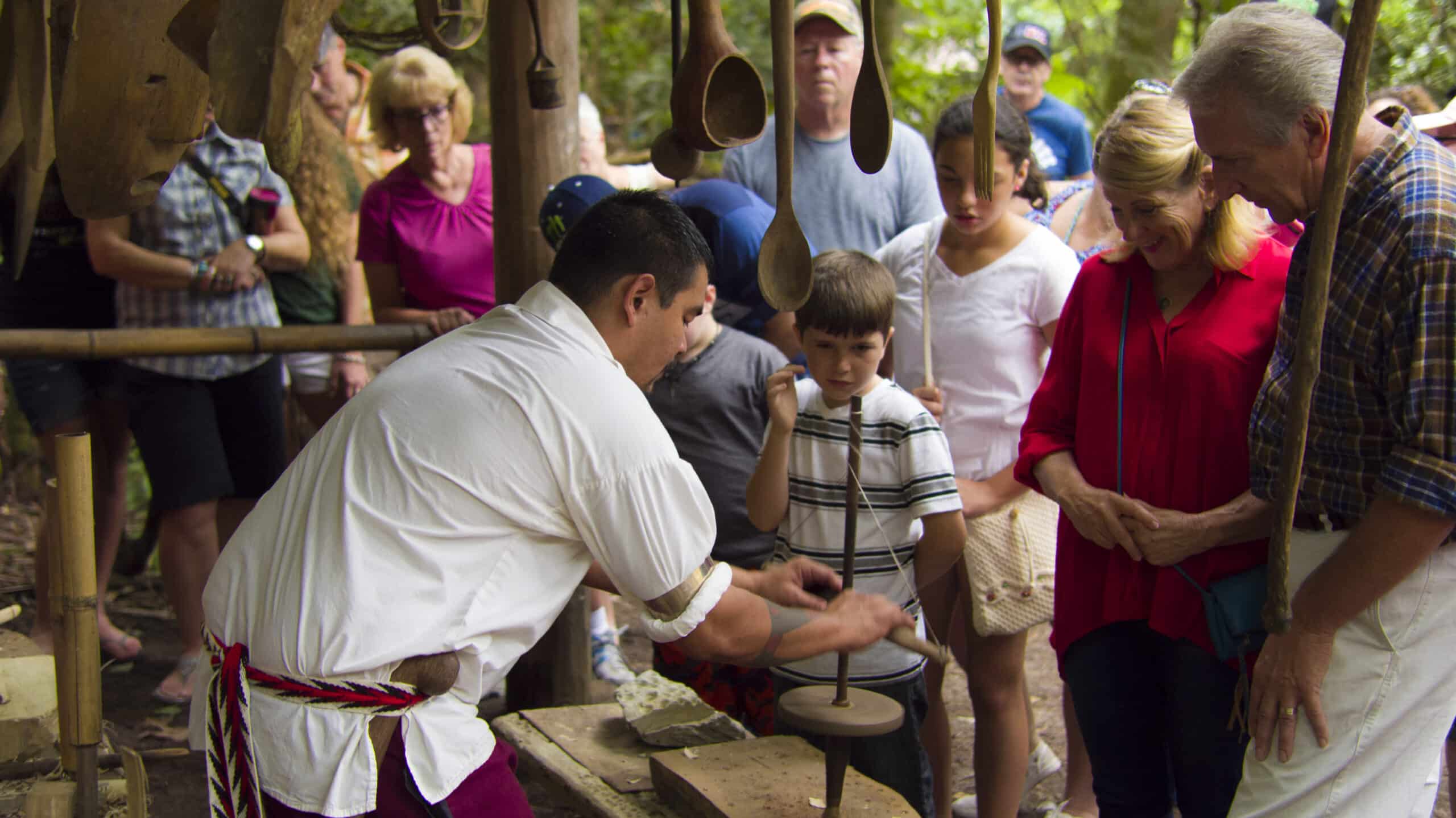 A craftsman demonstrates traditional pottery making to an interested group of people, including children and adults, at the Oconaluftee Indian Village in an outdoor setting surrounded by trees.
