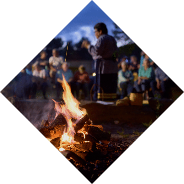 A diamond-shaped image of people gathering around a campfire at dusk, with a woman in sharp focus roasting a marshmallow, and others blurred in the background, conveying a sense of communal culture.