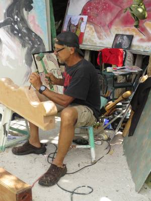 A man wearing sunglasses and a cap sits while painting on a canvas at his cluttered workspace, surrounded by various art supplies and paintings