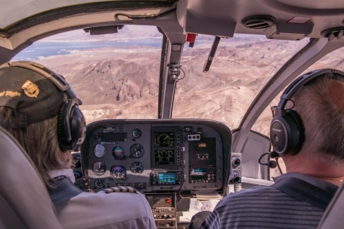 Two pilots in the cockpit of a Cherokee helicopter, viewing mountainous terrain through the windshield, with focus on the instrument panel and scenery ahead.