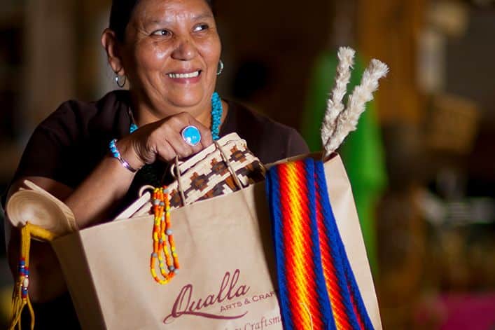 A person holds shopping bags from 'Qualla Arts and Crafts,' showcasing vibrant, multicolored beaded necklaces and a bracelet, implying a focus on indigenous crafts.