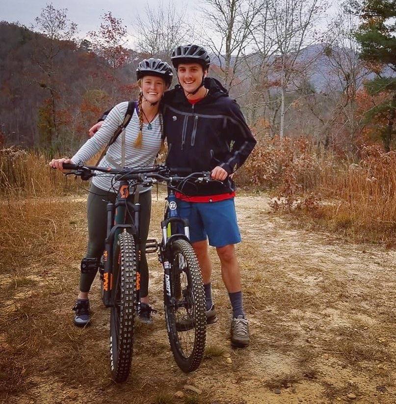 Two happy mountain bikers stand on a trail with their bikes, surrounded by autumn foliage under a cloudy sky. They are dressed in casual cycling gear and helmets, enjoying one of the many things to do